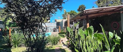 A garden pathway leading to a building with a pool, surrounded by lush greenery and a clear blue sky.