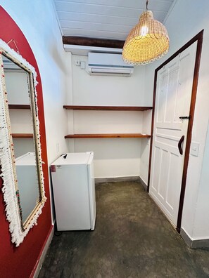 A compact room with a white refrigerator, a wall-mounted air conditioner, a wooden shelf, a mirror with a decorative frame, and a hanging light fixture. - Pousada Refugá (Porto Seguro)