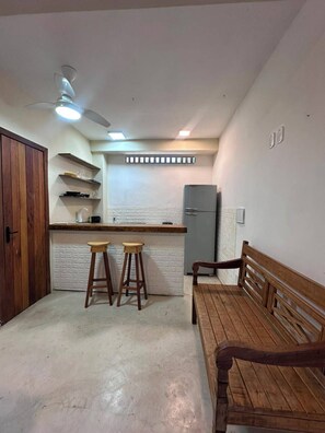 A compact kitchen with a wooden bench, two stools, and a refrigerator. - Pousada Refugá (Porto Seguro)