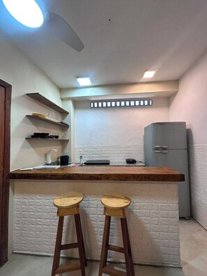 A compact kitchen with a wooden bar counter, two stools, a refrigerator, and built-in shelves. - Pousada Refugá (Porto Seguro)