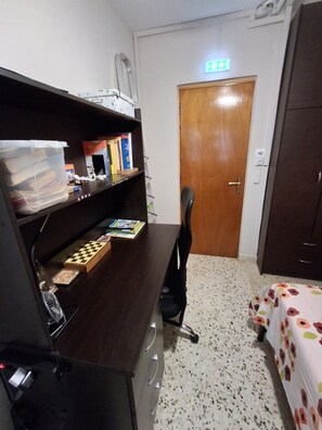 Ein kleines Hotelzimmer mit einem Holztisch, einem Stuhl, einem Bett mit blumigem Überwurf, einer Tür mit Notausgangsschild und einem an der Wand befestigten Regal mit Büchern und einem weißen Koffer.