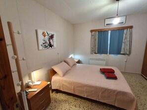 Ein Schlafzimmer mit Bett, Nachttisch, Lampe, Klimaanlage, einem Fenster mit Vorhängen und einem Wandbild.
