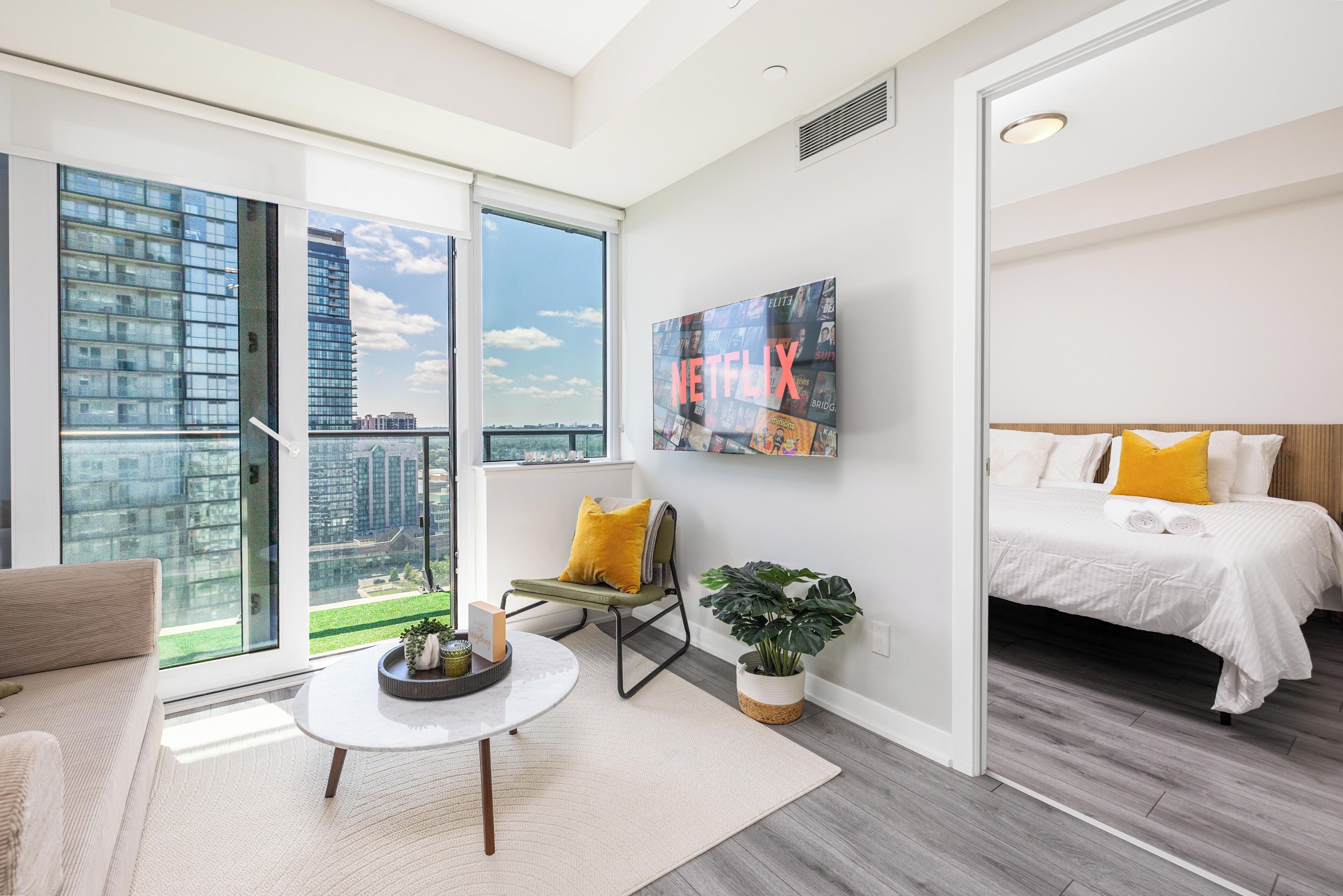 A modern hotel room with a large window, a bed with yellow pillows, a small table, and a wall-mounted TV.