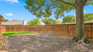 Un patio trasero con valla de madera, un árbol y una casa al fondo.