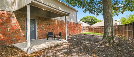A covered patio with two chairs, a brick wall, and a tree.
