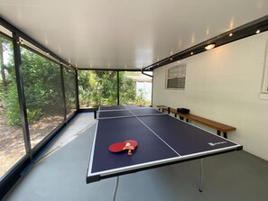 A ping pong table with a net in a room with large windows, a bench, and a wall-mounted picture. - Incredible 5-bedroom POOL Home Next Universal/Parks (Orlando)