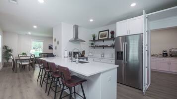 Cozinha moderna com ilha central, banquetas, geladeira de aço inoxidável e área de jantar com mesa e cadeiras.