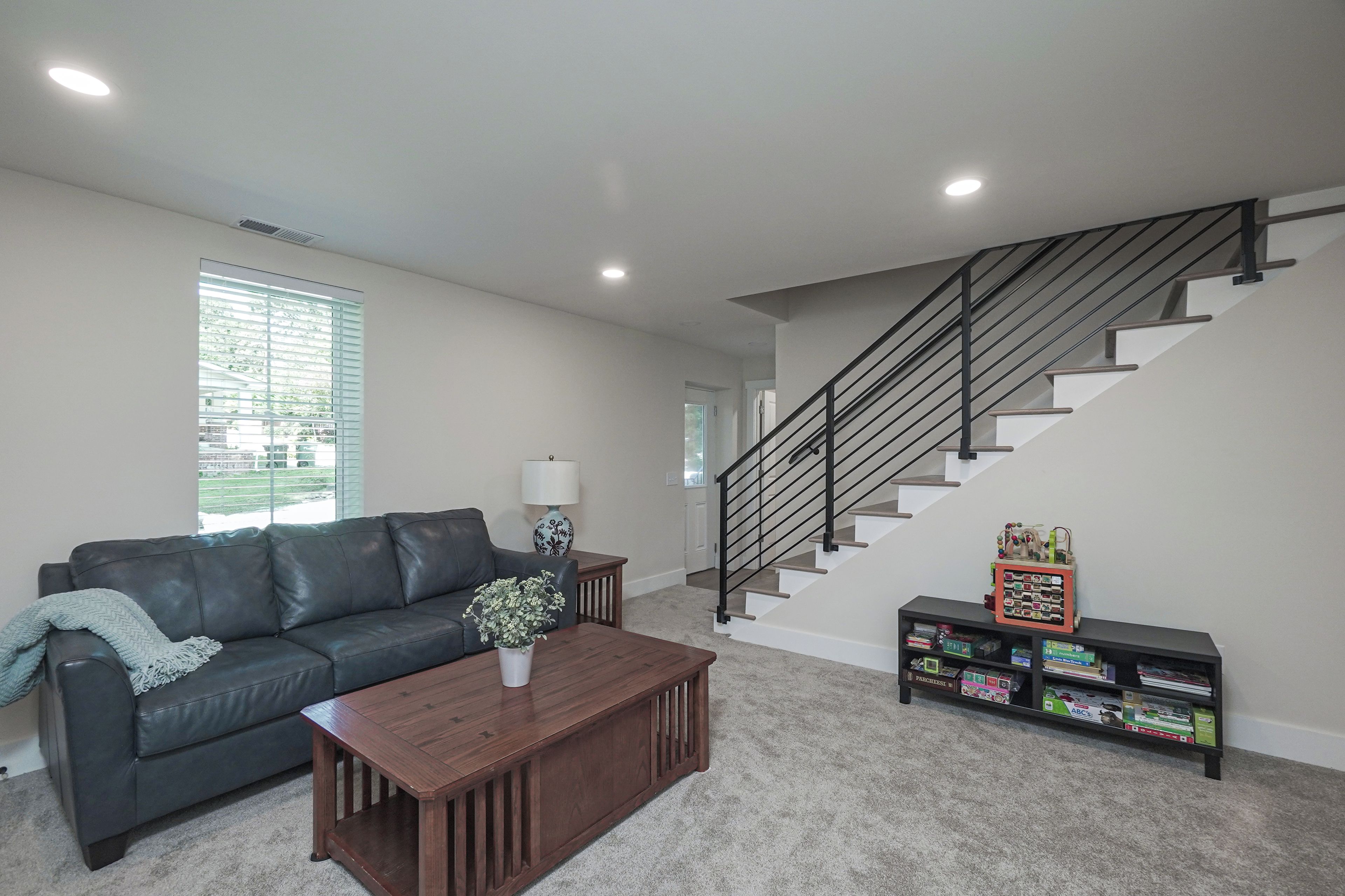 A living room with a dark leather sofa, a wooden coffee table, a potted plant, a bookshelf with children's books, and a staircase with a black railing.