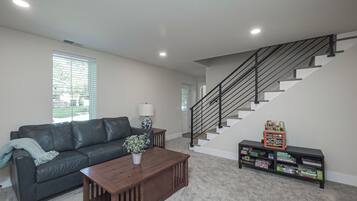 A living room with a dark leather sofa, a wooden coffee table, a potted plant, a bookshelf with children's books, and a staircase with a black railing.