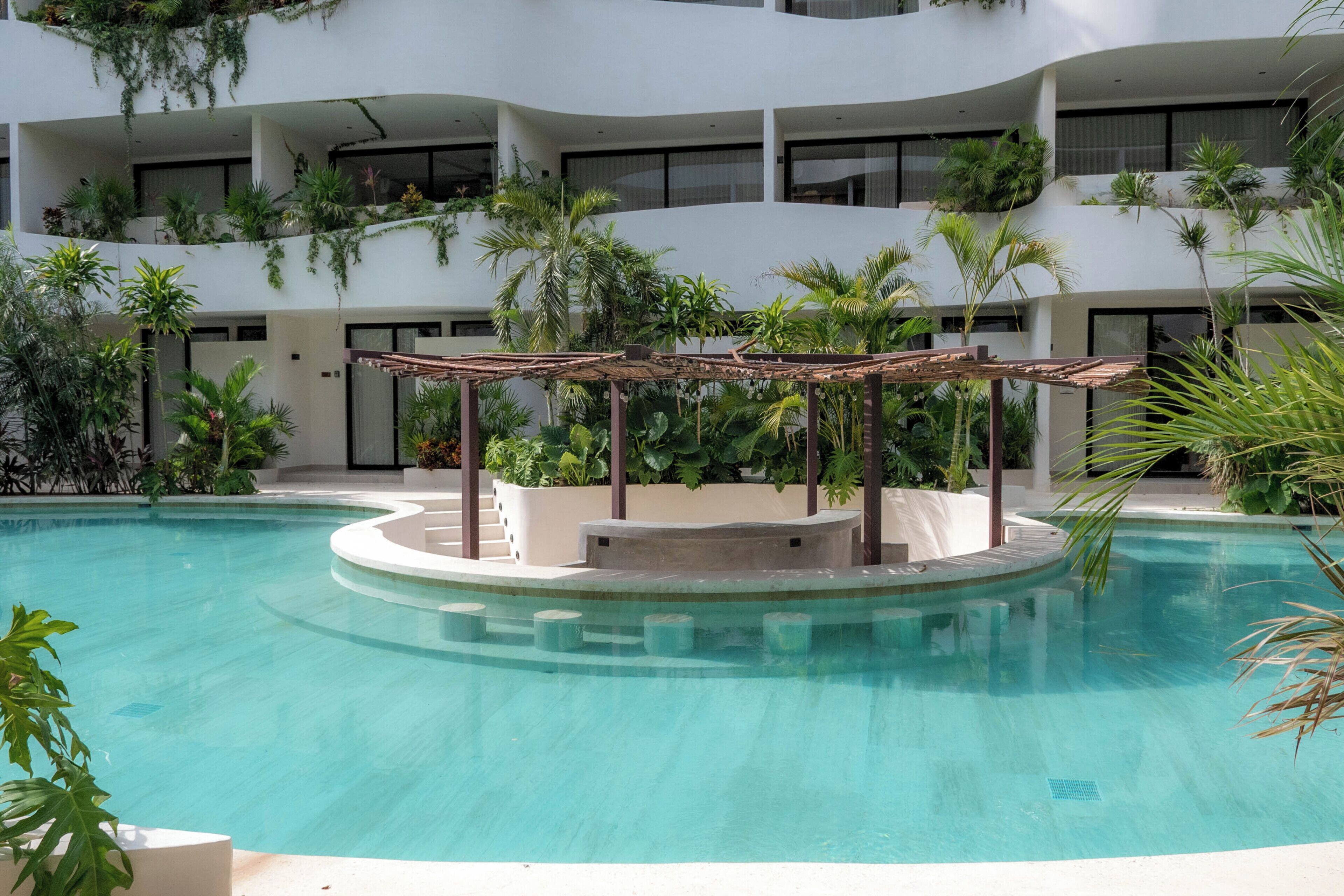 Ein modernes Hotel mit einem zentral gelegenen Schwimmbad, umgeben von üppiger Bepflanzung und zahlreichen Balkonen mit Pflanzen.