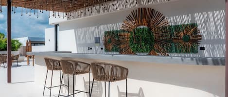 En utomhusbar med modern design, med en vit vägg som pryds av en dekorativ väggmålning i grönt och orange, samt tre barstolar i rotting.