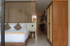 Ein Hotelzimmer mit einem großen Bett, einem kleinen Hocker, einem Holzschrank und einer Küchenzeile mit einer Pendelleuchte.
