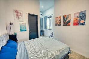 A bedroom with a bed, a desk, a chair, and a window. There are wall-mounted air conditioning units and artwork on the walls.