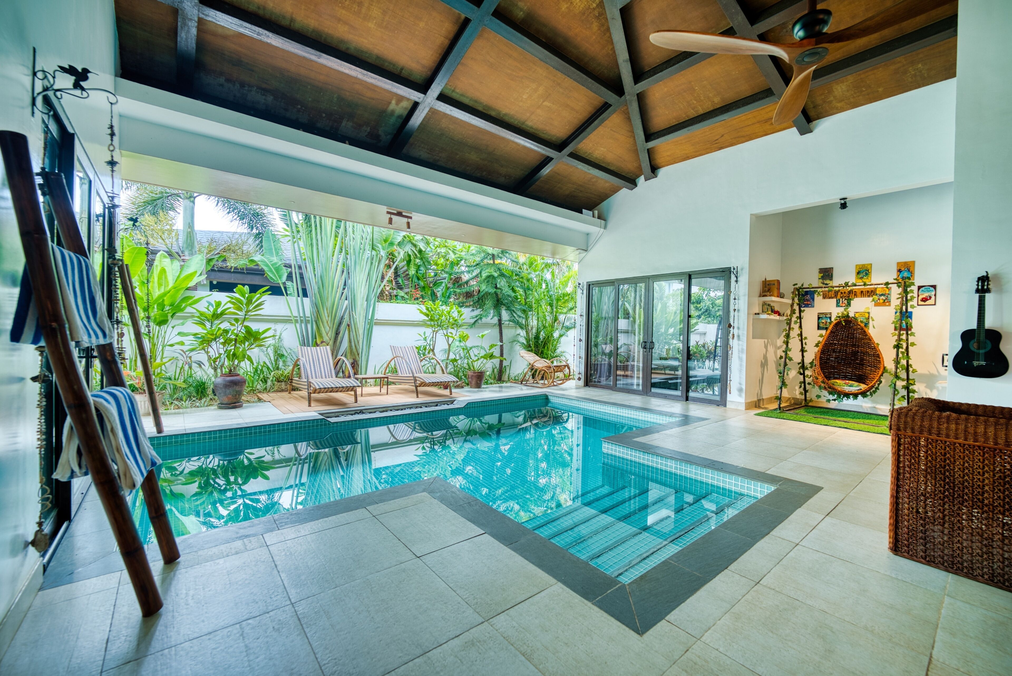 A modern indoor swimming pool with clear glass walls, surrounded by tropical plants and furnished with lounge chairs.