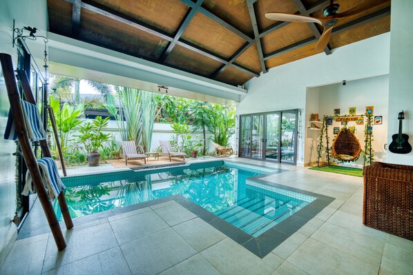 A modern indoor swimming pool with clear glass walls, surrounded by tropical plants and furnished with lounge chairs.
