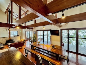 A spacious room with a large wooden table, benches, a fireplace, and large windows offering views of snow-covered trees. - Bears Den Hakuba | Private Lodge | 5 Bedrooms with Private Bathrooms, Hot Tub, and Walking Distance to the Ski Resort (白馬村)