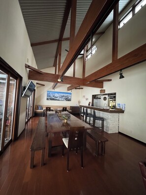 A spacious dining area with a long wooden table and benches, a kitchen area with a bar, and a large window providing natural light. - Bears Den Hakuba | Private Lodge | 5 Bedrooms with Private Bathrooms, Hot Tub, and Walking Distance to the Ski Resort (白馬村)