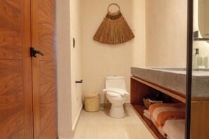 A bathroom with a wooden door, a toilet, a woven wall hanging, and a shelf with towels. - Vrbo Property (Tulum)