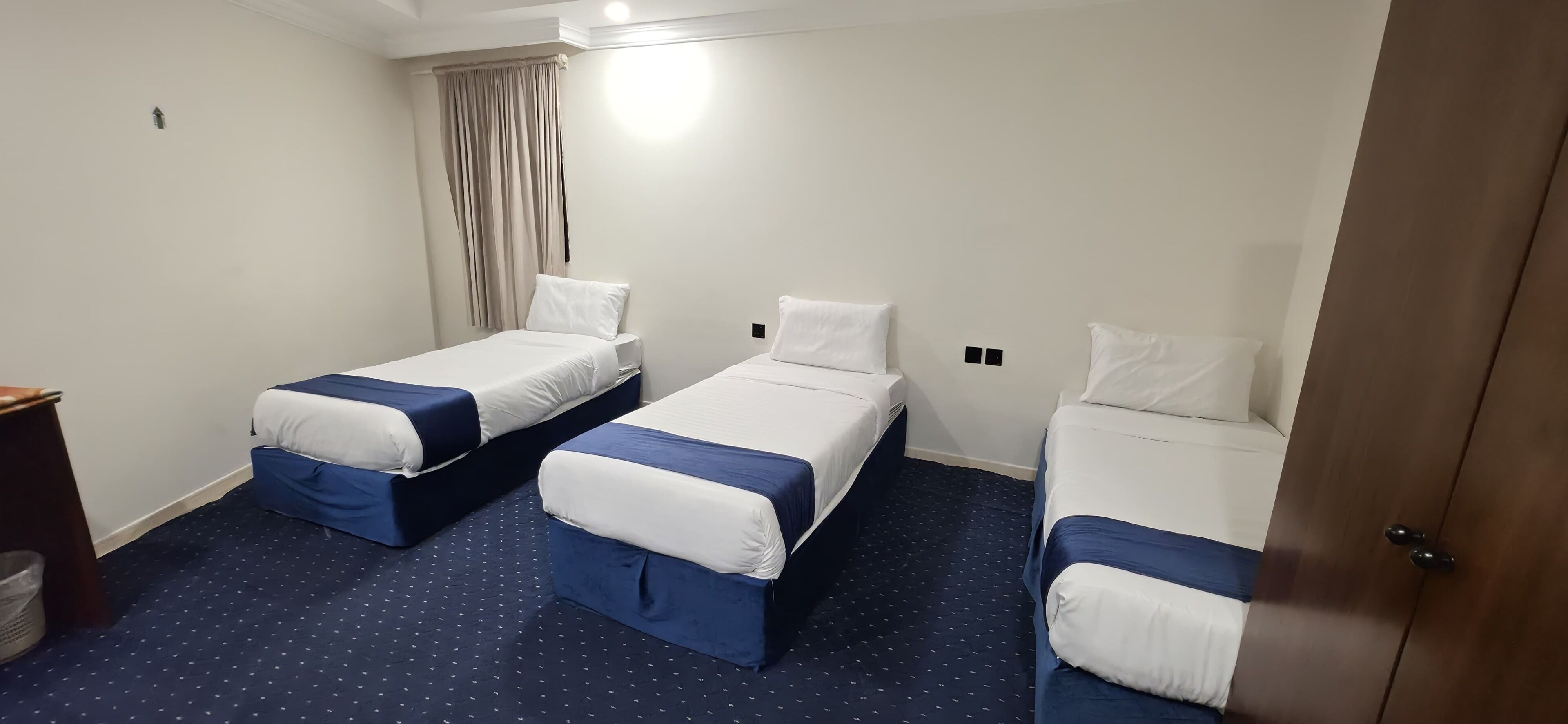 A hotel room with three single beds, each with a blue and white bedspread, a small bedside table, and a waste bin.