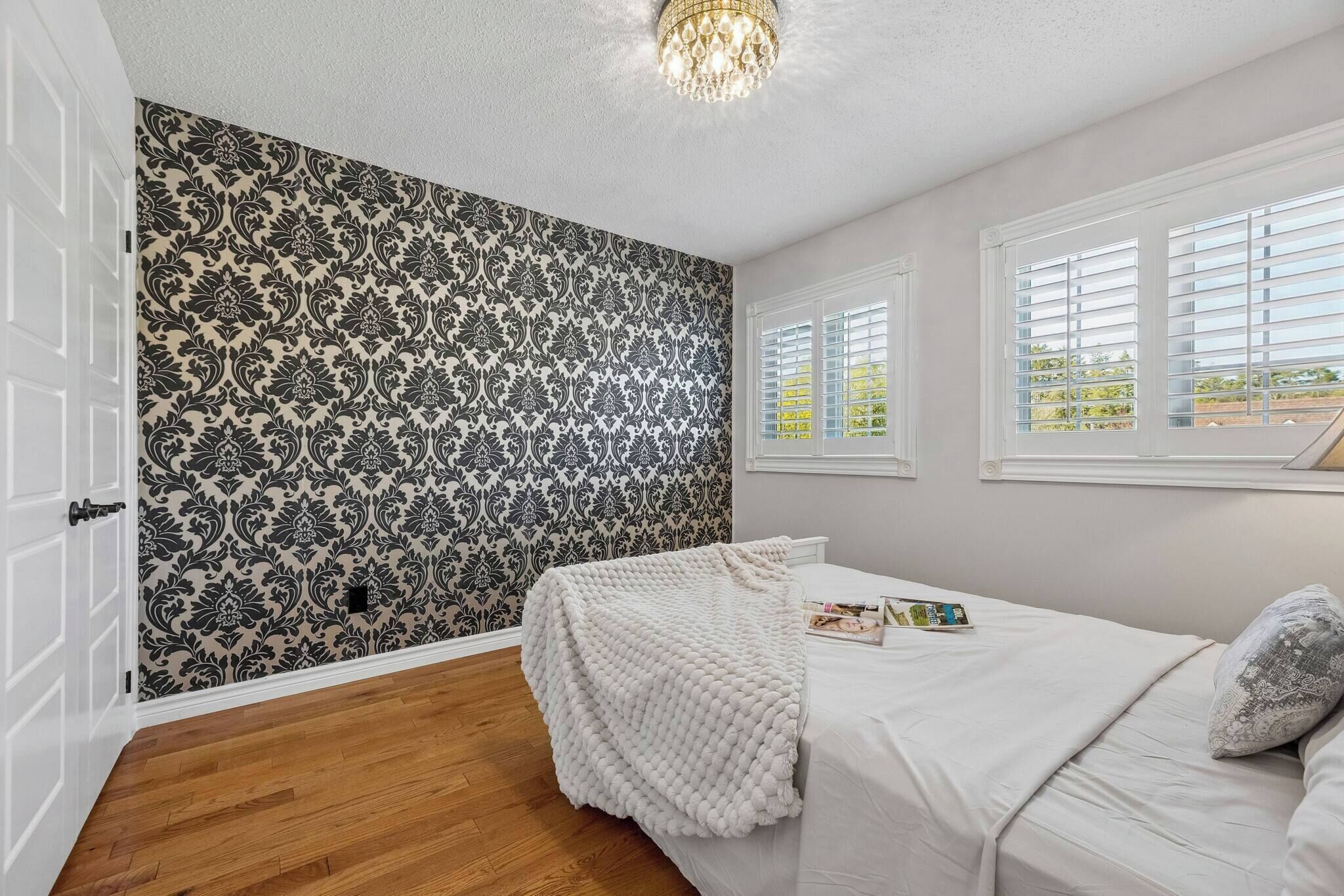 A bedroom with a patterned wallpaper, a bed with a white comforter, a white throw blanket, and a magazine on the bed.