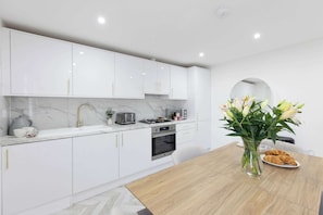 A modern kitchen with white cabinets, a marble countertop, and a wooden dining table with a vase of flowers. - Stylish Home Near Chelsfield Station, Free WiFi (Orpington)