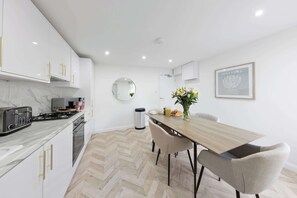 A modern kitchen with white cabinets, a marble countertop, and a dining area with a wooden table and chairs. - Stylish Home Near Chelsfield Station, Free WiFi (Orpington)