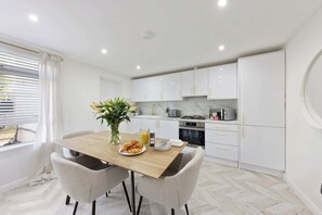 A modern kitchen with white cabinets, a dining table with chairs, and a window with blinds. - Stylish Home Near Chelsfield Station, Free WiFi (Orpington)