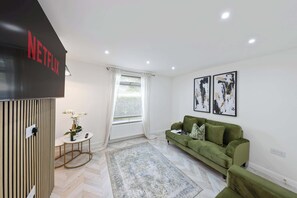 A modern living room with a green sofa, a small table with a bottle and glasses, a large window with curtains, and two abstract paintings on the wall. - Stylish Home Near Chelsfield Station, Free WiFi (Orpington)