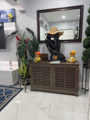 A reception area with a large mirror, a plant, a cabinet with figurines, and a wall-mounted sign. -  Beautiful and updated Cozy 2 Bedroom,2 Bathroom inPeaceful Portmore,Sleeps six. (Portmore)