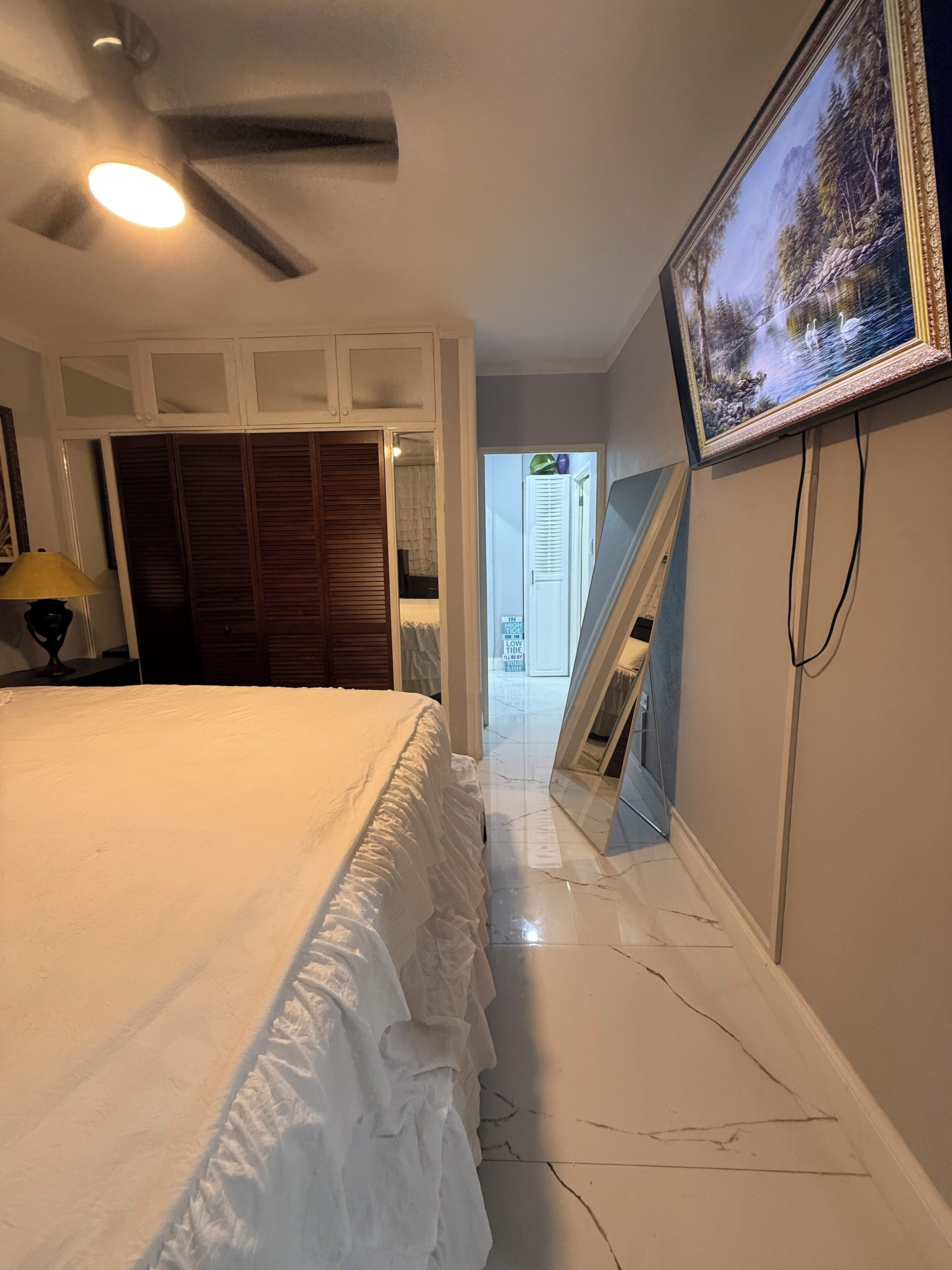 Un dormitorio con cama, ventilador de techo, televisor montado en la pared y una puerta que da al baño.