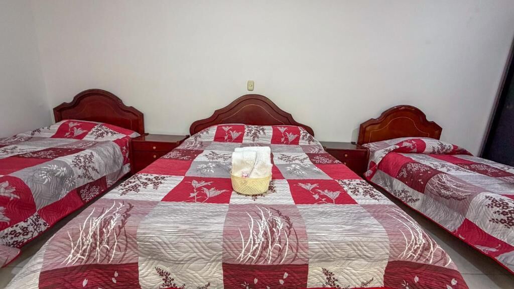 A hotel room with three beds, each with a wooden headboard and a floral-patterned bedspread. A basket with towels is placed on the floor between the beds.