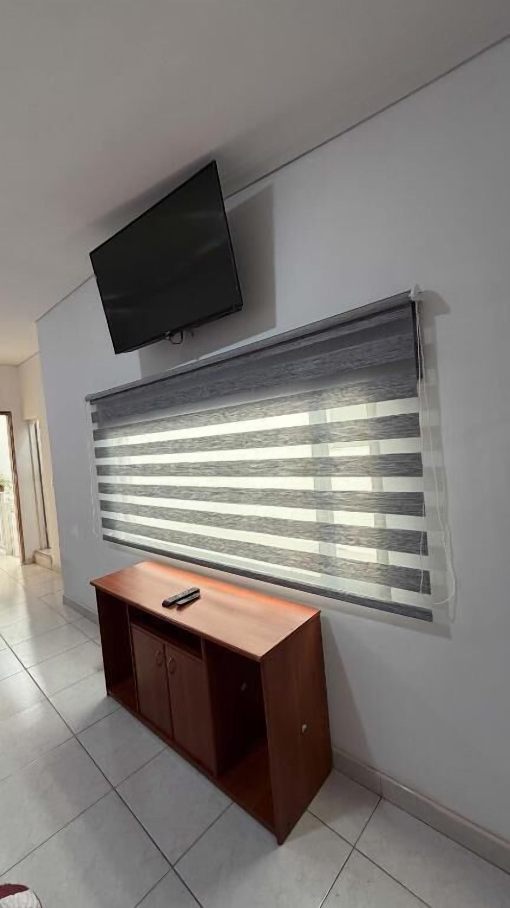 Une chambre dotée d’un téléviseur à écran plat fixé au mur, d’un bureau en bois avec une télécommande, et d’une fenêtre avec des stores horizontaux.