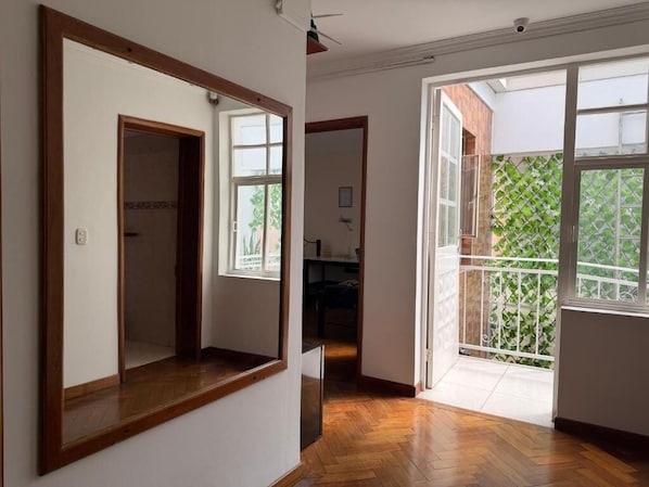 Un pasillo con suelo de madera, una puerta de armario espejada y un cerramiento de vidrio corredizo que da a un balcón con valla de celosía verde.