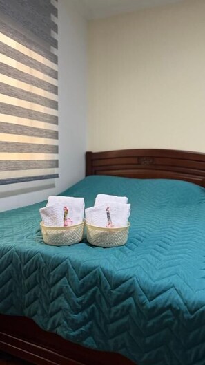 Una cama con colcha color turquesa, dos toallas blancas en cestas y una ventana con persianas horizontales.