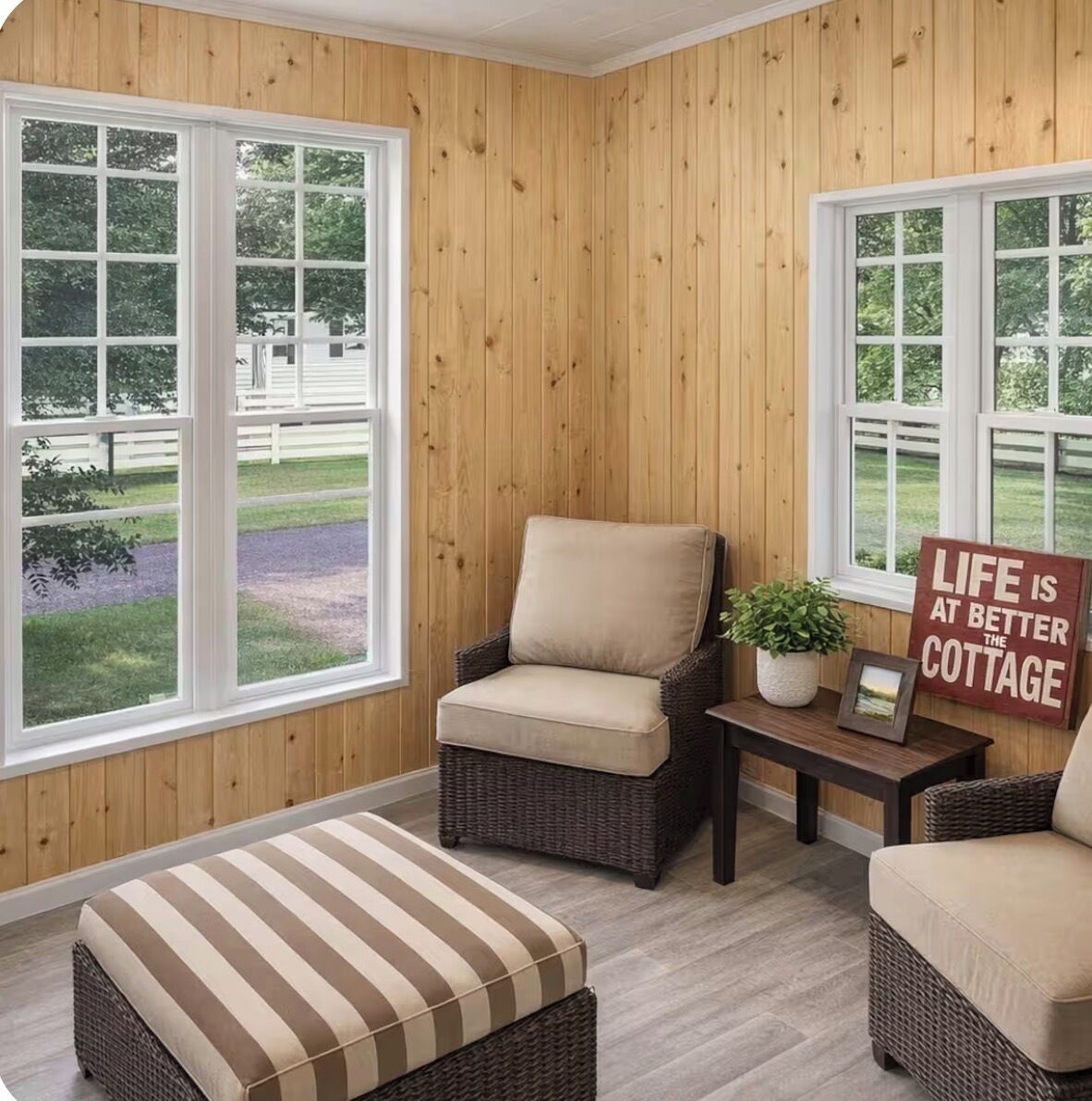 A cozy room with wooden paneled walls, a wicker chair, a cushioned ottoman, and a small table with a potted plant and a sign.