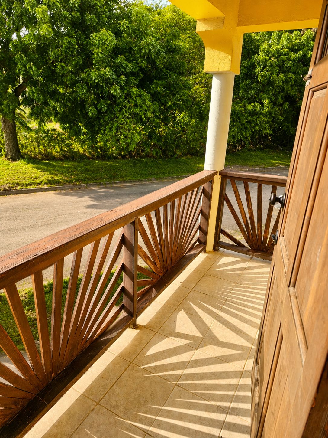 Un balcon en bois avec une balustrade ornée, un sol carrelé et une vue sur une route et la verdure.