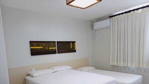 Et hotelværelse med to senge, en fastgjort aircondition og et vindue med gardiner.
