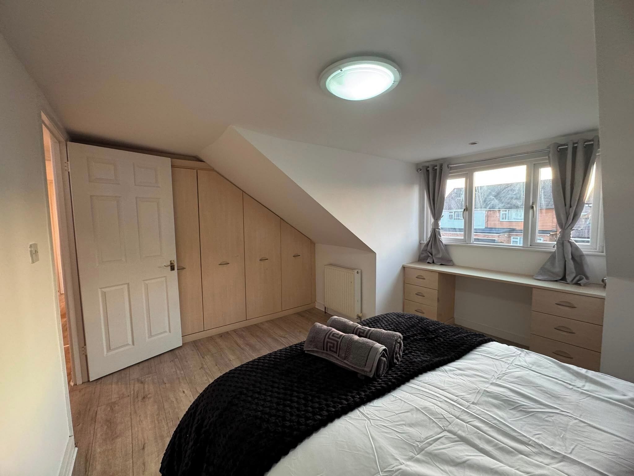 A bedroom with a sloped ceiling, a bed with a black and white duvet, a window with curtains, a radiator, and a wooden wardrobe.