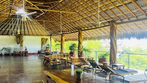 An outdoor dining area with wooden tables and chairs, potted plants, and a thatched roof structure.