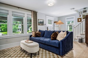 A living room with a blue sofa, a patterned rug, and a view of a green landscape through large windows. - NEW! Prelaunch Pricing Close to Track and Downtown (Saratoga Springs)