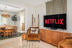 Ein Wohnzimmer mit Fernseher, der 'NETFLIX' anzeigt, einem Holzschrank, einem braunen Ledersessel und einem Essbereich mit Holztisch und Stühlen.