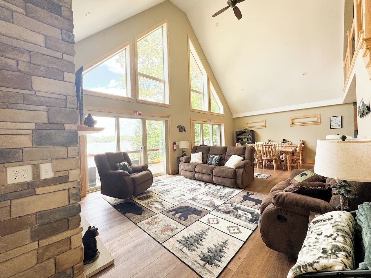 A spacious living room with a stone fireplace, large windows, and a high ceiling fan.
