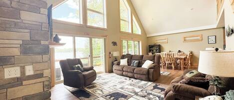 A spacious living room with a stone fireplace, large windows, and a high ceiling fan.