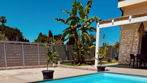 Una piscina azul cristalina con borde blanco, rodeada por un patio con sillas y una mesa, y una palmera al fondo.