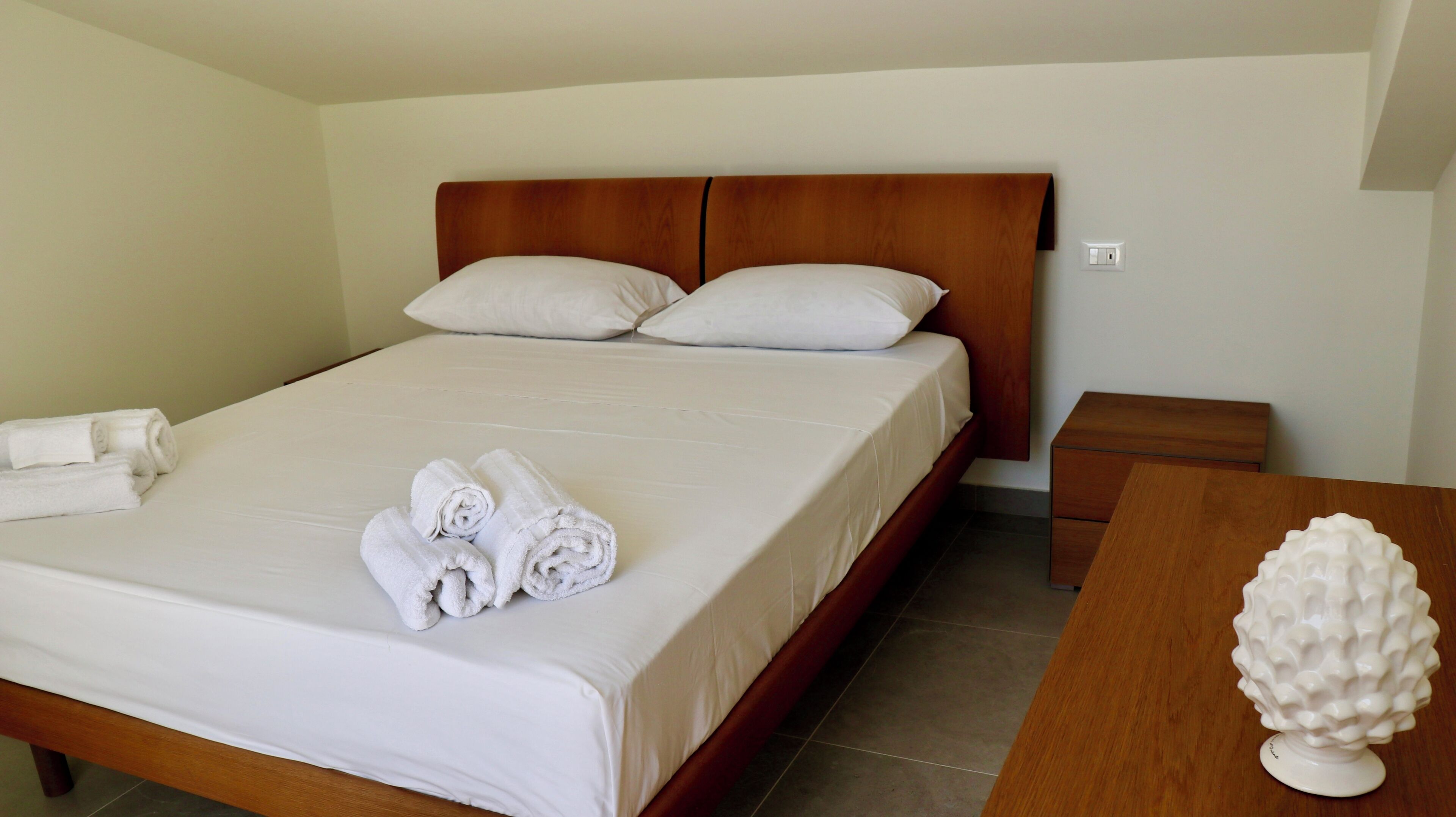 Una cama bien hecha con sábanas blancas y toallas enrolladas, un cabecero de madera, una mesita de noche con un objeto decorativo blanco, y una pequeña mesita auxiliar de madera.