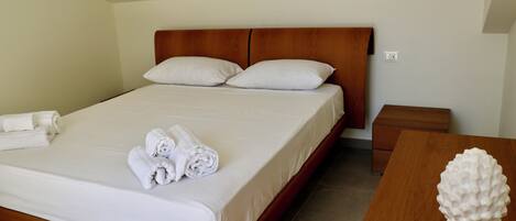 Una cama bien hecha con sábanas blancas y toallas enrolladas, un cabecero de madera, una mesita de noche con un objeto decorativo blanco, y una pequeña mesita auxiliar de madera.