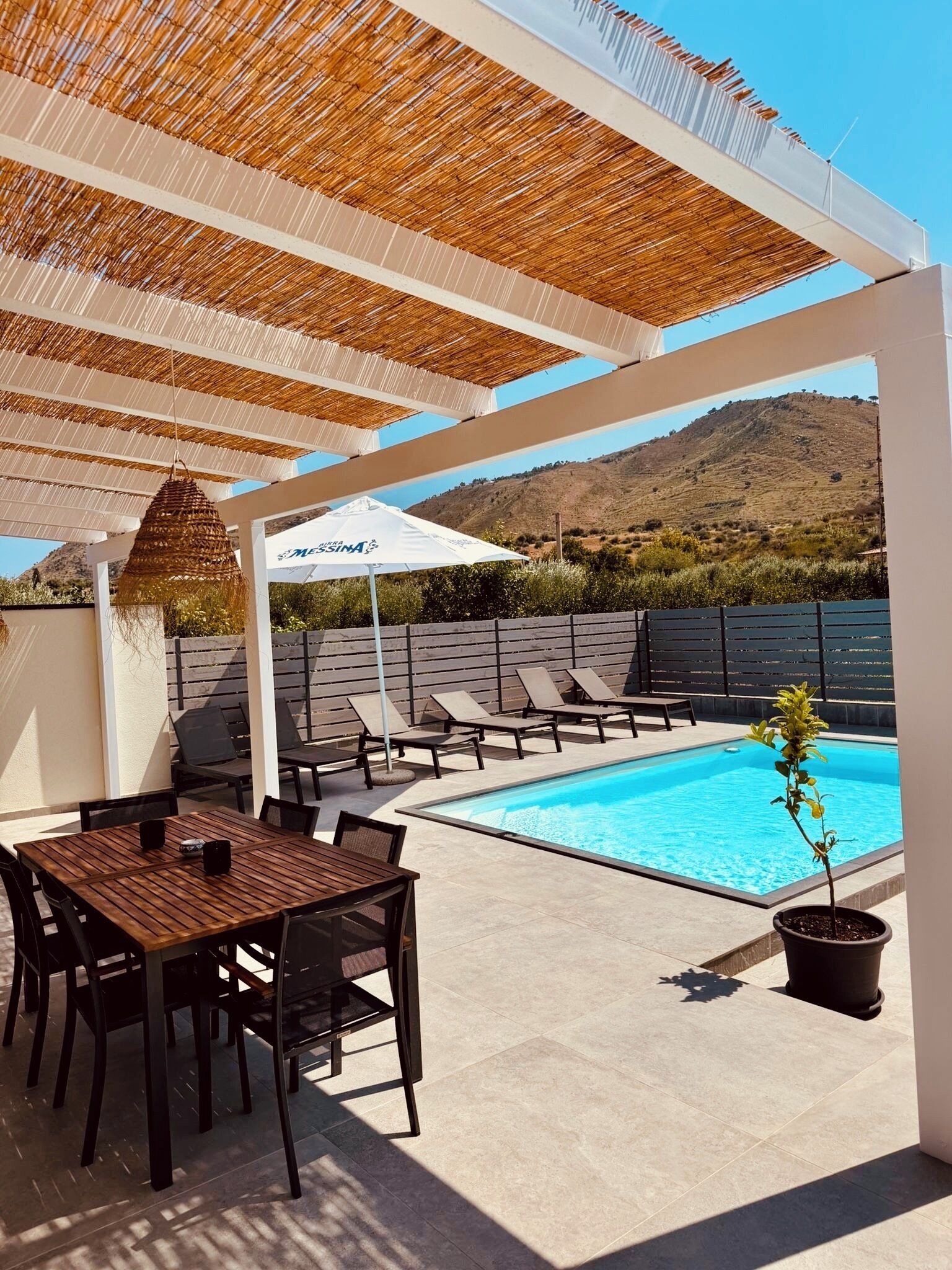 A poolside area with a table and chairs, a hanging umbrella, and a view of a mountainous landscape.