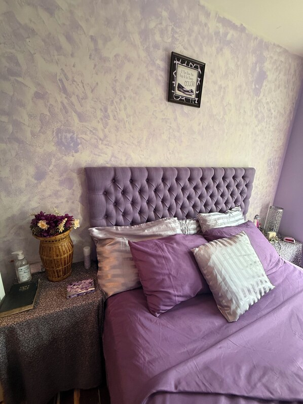 Un dormitorio con cabecera capitonada, cama con ropa de cama púrpura, mesita de noche con un jarrón de flores y una cita enmarcada en la pared.