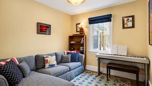Een woonkamer met een grijze bank, een piano, een boekenkast en een raam met uitzicht.