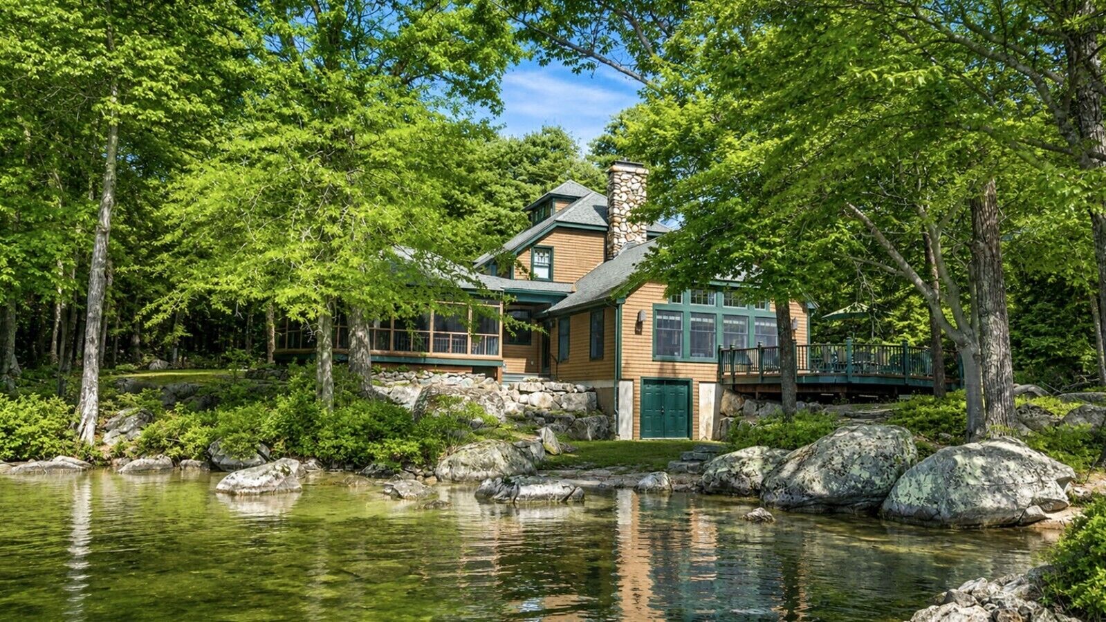A lodge with a stone chimney, wooden deck, and a pond with rocks.
